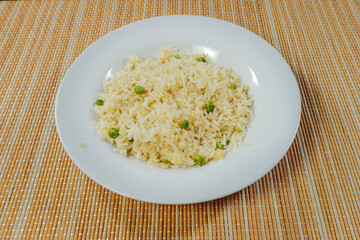 Cooked jasmine rice with green peas on a white plate and bamboo table surface.