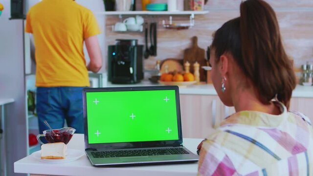 Business woman working on laptop with green screen in kitchen and boyfriend is cooking morning meal. Green screen chroma mock up isolated mockup background ready to be replaced with your text, logo or