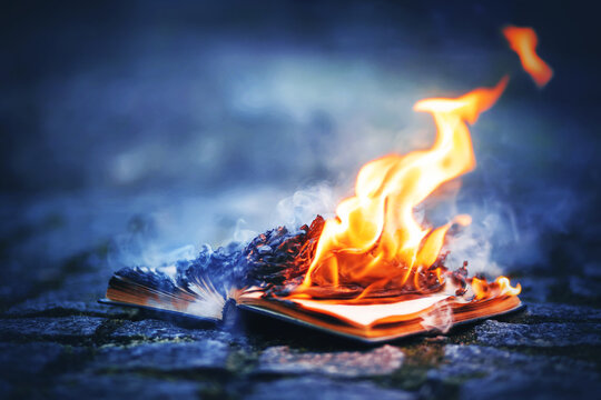 An old book lies on a rocky path made of cobblestones and its pages burn with a bright strong flame and smoke blue smoke at night. Magic. Censorship.