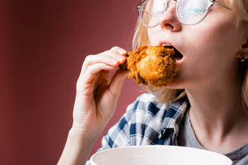 Closeup portrait of a blonde girl who brings a chicken leg to her mouth while holding a bucket of...