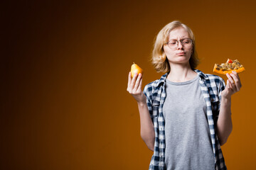 The blonde girl in glasses holds a pear and a slice of pizza in her hands thinking what to eat. The end of a healthy and unhealthy diet on an orange background.