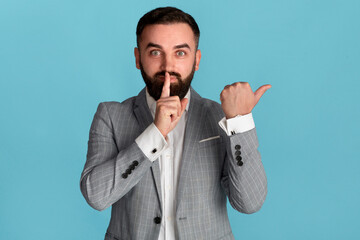 Attractive entrepreneur showing HUSH gesture and pointing aside on blue background