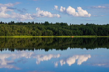 Fototapeta premium Beautiful summer landscape. View of the lake and forest. Red boat in the distance. Outdoor recreation and fishing on the lake. Clouds are reflected in the water. Travel and vacation in the countryside