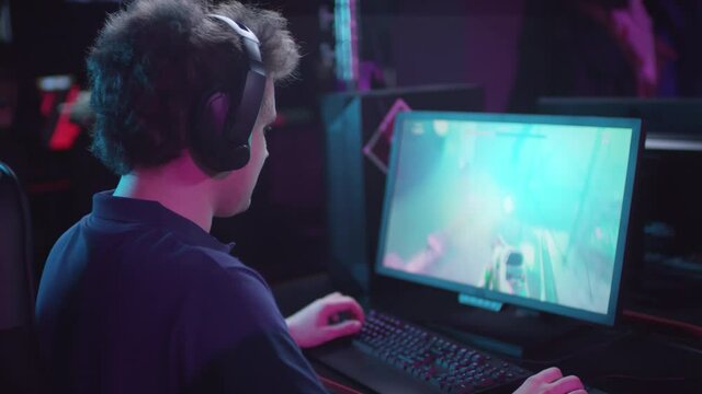 Sequence of shots footage of young man with curly hair wearing headphones with microphone playing shooter video game and failing it