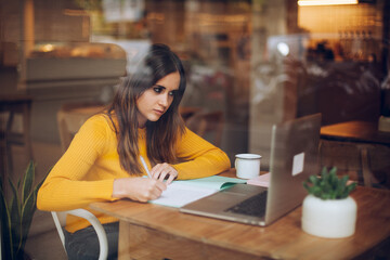 Positive caucasian female checking news from networks studying in coffee shop, girl freelancer finding  information on web page writing notes with a cup of tea