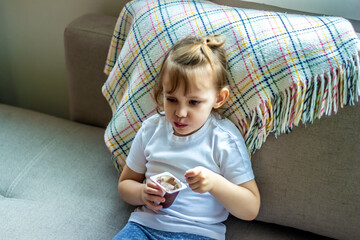 Top view of cheerful girl sitting on sofa at home and eating tasty pudding. Pretty little child looking at camera, smiling and posing. Concept of enjoyment and lunch.