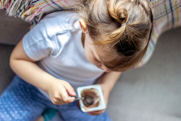 Top view of the cute girl eating her favorite chocolate dessert. Beautiful little girl enjoying her moment. Toddler in white tshirt sitting on bed alone and eating afternoon snack.