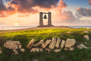 Picturesque landscape with an ancient foggy bell in the center of the frame. Spring evening by the sea, magical sunset, ancient masonry, fresh grass and foggy bell. Copy space on the colorful sky.