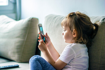 Beautiful little girl playing game or watching video on smartphone mobile.Girl watching cartoons or browsing internet. Side view portrait of little girl using smartphone while sitting on sofa.
