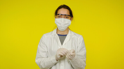 Young doctor in protective medical mask and glasses removes latex gloves from hands. Nurse in a white coat on a yellow background after the patient