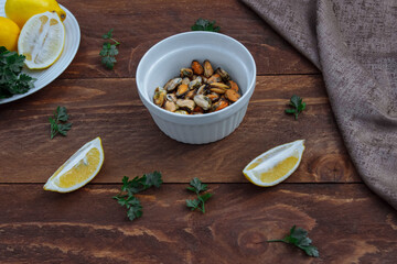 Peeled pickled mussels in a white bowl with slices and whole lemons and a bunch of parsley on a white plate with a brown napkin on a wooden background