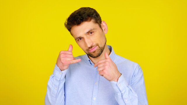 Call me. Handsome bearded man flirts and asks to call him back. Guy in light blue shirt on a yellow background with copy space imitates phone with fingers. Place for text or product