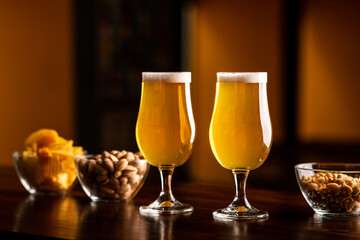 Two glasses with light beer and foam on wooden bar counter in interior of pub, pistachios, crackers and nuts in glass plates nearby