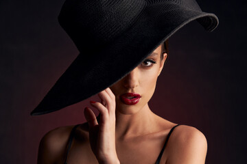 portrait of beautiful tanned girl with professional makeup, red lips, on a burgundy background in a black dress with straps and black hat that covers her eye she looks at the camera with serious face