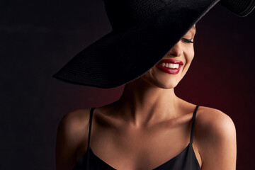 portrait of a beautiful tanned girl with professional makeup on a burgundy background in a black dress with straps, black hat that covers her eye, she smiles with white teeth