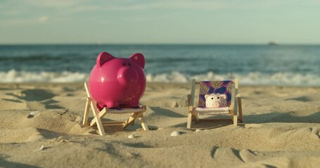 Two piggy banks sit on mini sun loungers on the sandy beach. A man's hand throws a coin into a piggy bank. Male Hand Throws a Coin Into a Piggy Bank a Pig on a Sandy Shore. Slow Motion. - Powered by Adobe