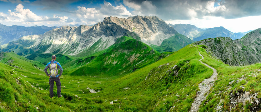 Hiker Man on Hiking Trail with Backpack standing relaxed and enjoying panoramic view to mountains. Alps, Hohe Gaenge, Allgau, Bad Hindelang, Hinterstein, Bavaria, Germany.