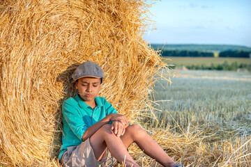 One young boy sits at a haystack in a field on a sunny day and thinks about something.