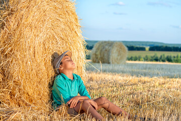 Naklejka na ściany i meble One young boy sitting with his eyes closed at a haystack in a field on a sunny day and dreaming of something.