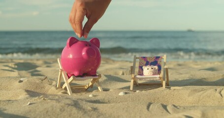 Two piggy banks sit on mini sun loungers on the sandy beach. A man's hand throws a coin into a piggy bank. Male Hand Throws a Coin Into a Piggy Bank a Pig on a Sandy Shore. Slow Motion. - Powered by Adobe