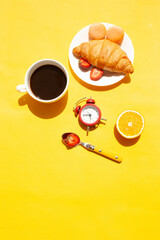 Cup of coffee, red alarm clock, croissant and orange. Morning breakfast on a yellow background. Sunny day