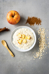Warmes Frühstück mit Porridge und Apfel in einer weißen Schüssel. Draufsicht, Zutaten, Gewürze, Holzlöffel, grauer HIntergrund.