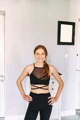 Midshot of a sporty smiling girl. She stands with her arms on her hips, looking and smiling into camera. Home workout, online training