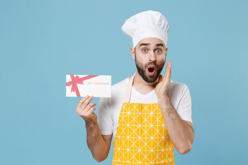 Shocked young bearded male chef or cook baker man in apron white t-shirt toque chefs hat isolated on blue background. Cooking food concept. Mock up copy space. Hold gift certificate put hand on cheek.