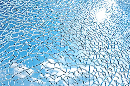 broken glass abstract texture background against sun backlit blue sky with clouds. Shattered thick window pattern. Destruction of windshield surface. Damage of bulletproof glass shield Design backdrop