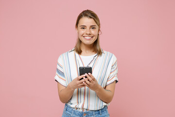 Smiling young blonde woman girl in casual striped shirt posing isolated on pastel pink background studio portrait. People lifestyle concept. Mock up copy space. Using mobile phone, typing sms message.