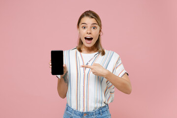 Shocked young blonde woman girl in casual striped shirt isolated on pastel pink background. People lifestyle concept. Mock up copy space. Pointing index finger on mobile phone with blank empty screen.