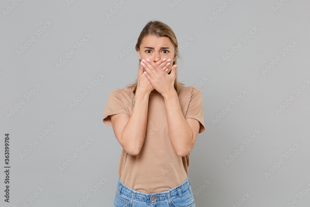 Poster Shocked worried young blonde woman girl in casual beige t-shirt posing isolated on gray background studio portrait. People emotions lifestyle concept. Mock up copy space. Covering mouth with hands.