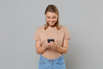 Smiling young blonde woman girl in casual beige t-shirt posing isolated on gray wall background studio portrait. People lifestyle concept. Mock up copy space. Using mobile phone, typing sms message.