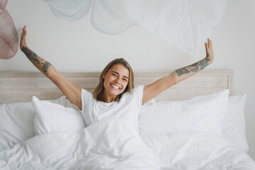 Smiling young girl in white t-shirt lying in bed with white sheet pillow blanket spending time in...