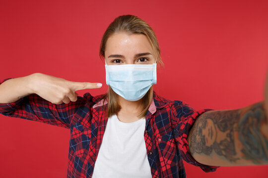 Close Up Girl In Checkered Shirt Isolated On Red Background. Epidemic Pandemic Coronavirus 2019-ncov Sars Covid-19 Flu Virus Concept. Doing Selfie Shot On Mobile Phone Point Index Finger On Face Mask.