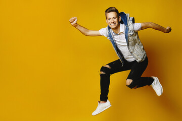 Fototapeta na wymiar Side view of happy young hipster guy in denim clothes jumping at the yellow background, isolated. Advertising mockup with copy space