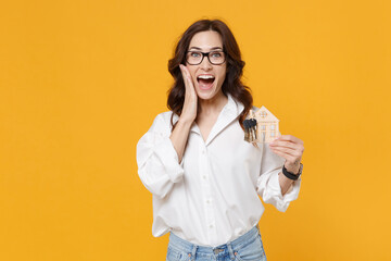 Excited young brunette business woman in white shirt glasses isolated on yellow background. Achievement career wealth business concept. Mock up copy space. Hold house bunch of keys, put hand on cheek.