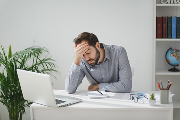 Exhausted tired business man in gray shirt sitting at desk work on laptop pc computer in light...
