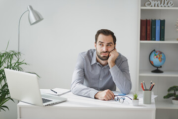 Boring young bearded business man in gray shirt sitting at desk working on laptop pc computer in...