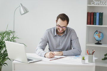 Smiling young bearded business man in gray shirt glasses sit at desk work on laptop pc computer in light office on white wall background. Achievement business career concept Writing notes in notebook.