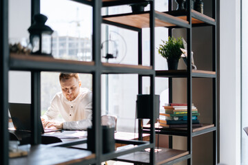 Serious young business man typing on laptop at office desk, background of large window. Concept of office working.