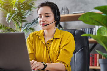 Beautiful freelancer female talking in online video conference using headset with microphone and laptop in home office desktop