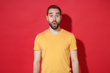 Shocked amazed young bearded man guy in casual yellow t-shirt posing isolated on red wall background studio portrait. People sincere emotions lifestyle concept. Mock up copy space. Keeping mouth open.