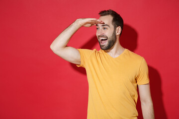 Fototapeta na wymiar Excited young bearded man guy in casual yellow t-shirt posing isolated on red wall background studio. People lifestyle concept. Mock up copy space. Holding hand at forehead looking far away distance.