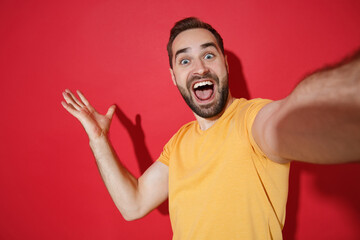 Fototapeta premium Close up of Shocked young bearded man guy in casual yellow t-shirt posing isolated on red background. People lifestyle concept. Mock up copy space. Doing selfie shot on mobile phone spreading hands.