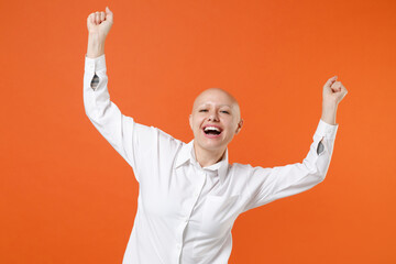 Fototapeta premium Joyful young bald business woman girl in white shirt posing isolated on orange background studio portrait. Achievement career wealth business concept. Mock up copy space. Clenching fists like winner.