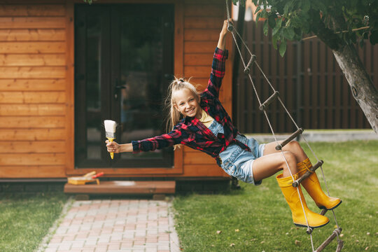 Beautiful teenage girl in denim overalls, a plaid shirt and yellow rubber boots, holding a brush in her hand at home, indulges and rides on a swing. Home Improvement, DIY Repair