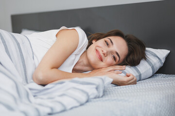 Obraz na płótnie Canvas Charming smiling young woman in white t-shirt sleeping lying in bed with striped sheet pillow blanket spending time in bedroom at home. Rest relax good mood lifestyle concept. Mock up copy space.