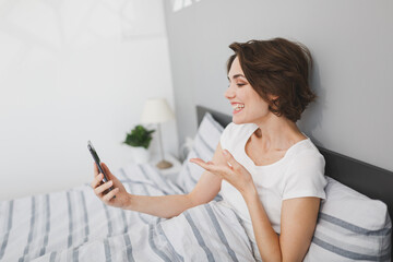 Cheerful smiling young brunette woman girl in white t-shirt lying in bed spending time in bedroom at home. Rest relax good mood lifestyle concept. Doing selfie shot on mobile phone, making video call.