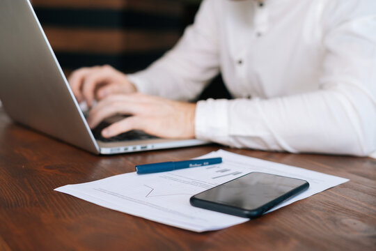 Close-up view of unrecognizable busy young businessman wearing fashion casual clothing is working on laptop computer at desk in modern office room ,mobile phone on table. Concept of office working.
